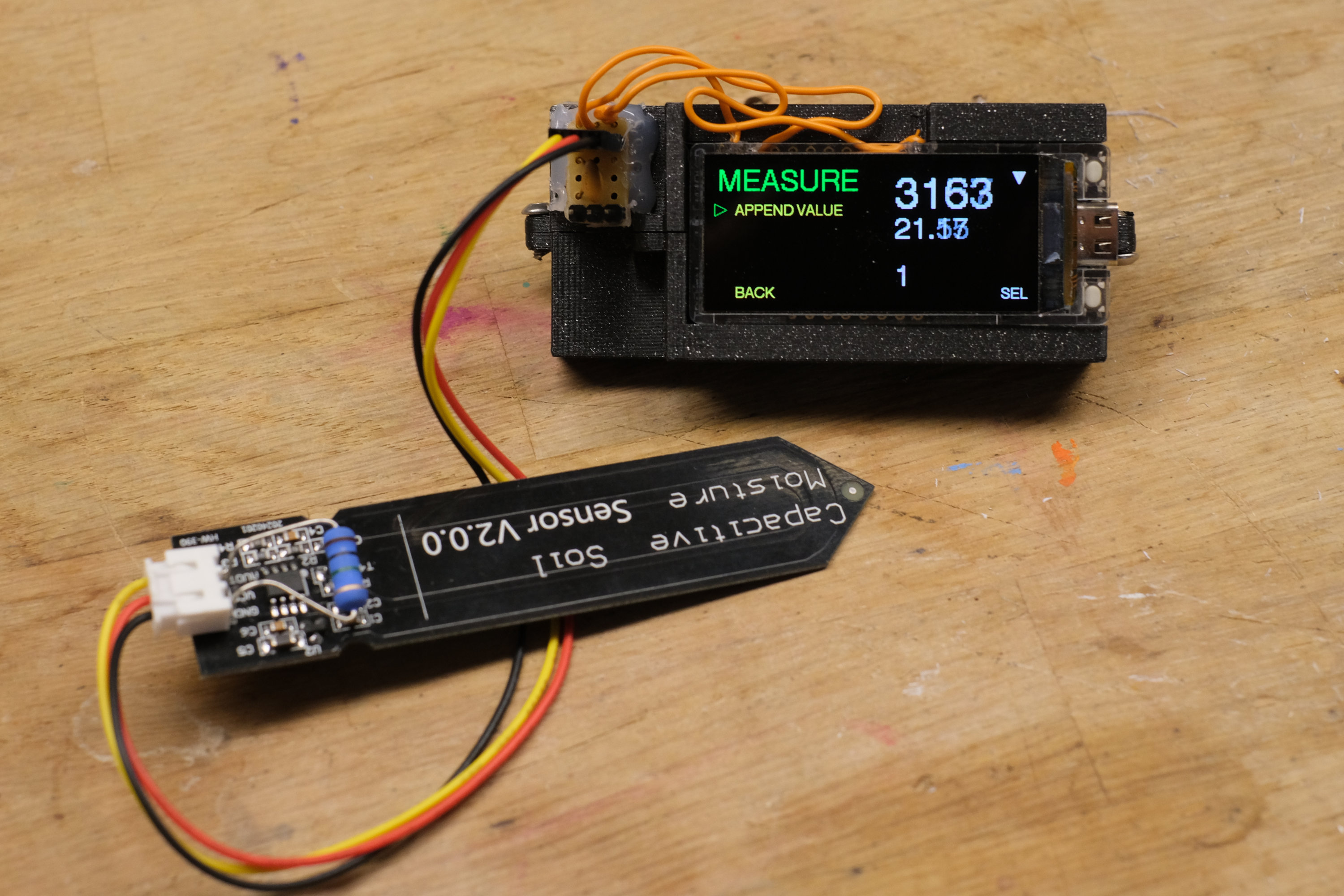 Ein Mikrocontroller mit Display in ein Gehäuse eingebettet. Daran angeschlossen ein kapatitiver Bodenfeuchtigskeitssensor